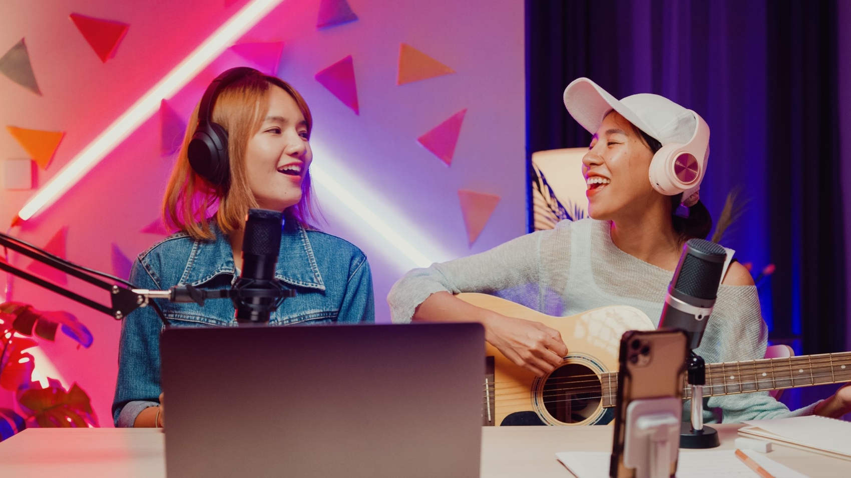 Happy Asian girl friends influencer wear headphone record podcast on laptop onair online live streaming play guitar sing song together for follower at home studio.
